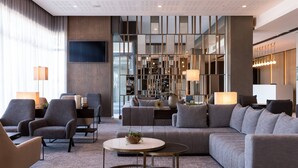 Um hall moderno de hotel com um sofá modular, poltronas, uma mesa de centro redonda e uma TV fixada na parede.