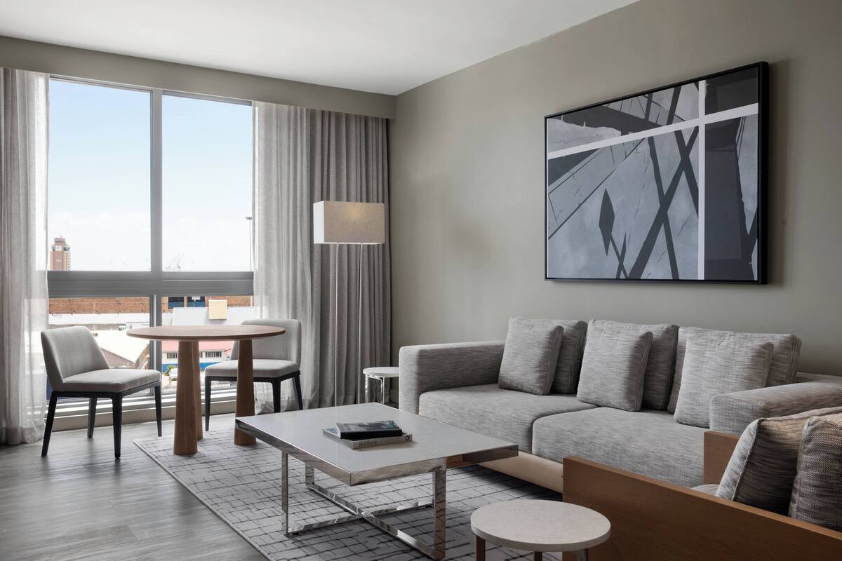 Moderni hotellihuone, jossa on harmaa sohva, pyöreä ruokapöytä ja suuri ikkuna, jossa on verhot.