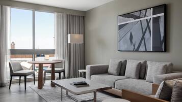 A modern hotel room with a grey sofa, a round dining table, and a large window with curtains.