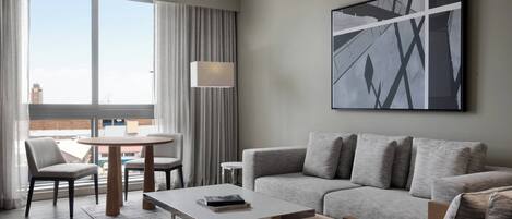 Una habitación de hotel moderna con un sofá gris, una mesa de comedor redonda y un ventanal con cortinas.