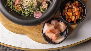 A bowl of hummus with garnishes, accompanied by slices of pickled vegetables and a dark bread on a wooden board.