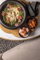 A bowl of hummus with garnishes, accompanied by slices of pickled vegetables and a dark bread on a wooden board.