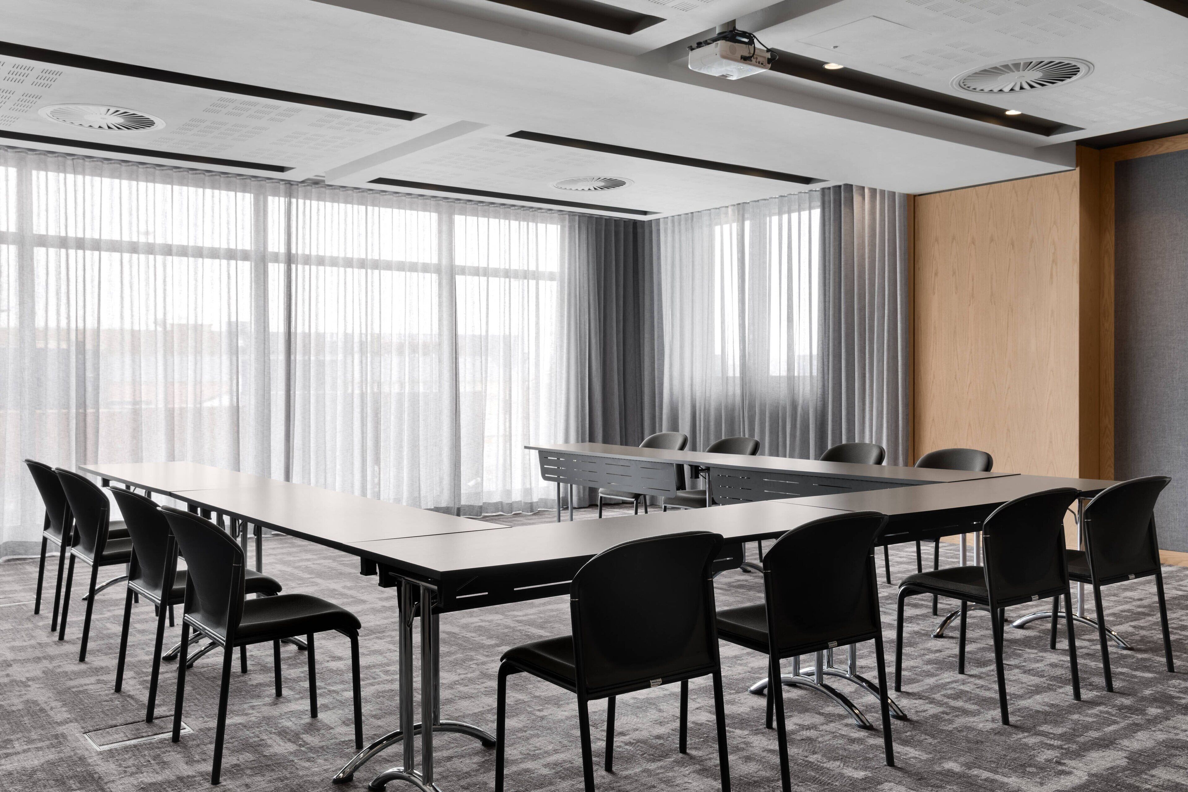 A conference room with a long table and multiple chairs, large windows with curtains, and a ceiling with recessed lighting and ventilation.