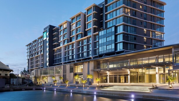 Um hotel de vários andares com design arquitetônico moderno, destacando uma entrada imponente com portas de vidro e uma área externa bem iluminada com degraus que levam à entrada.