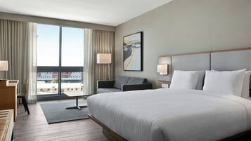 A hotel room with a large bed, a desk, a chair, and a view of the city through large windows.