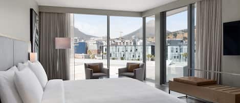 Una habitación de hotel moderna con una cama grande, dos sillones, un banco de madera pequeño y un ventanal con vistas a la ciudad.