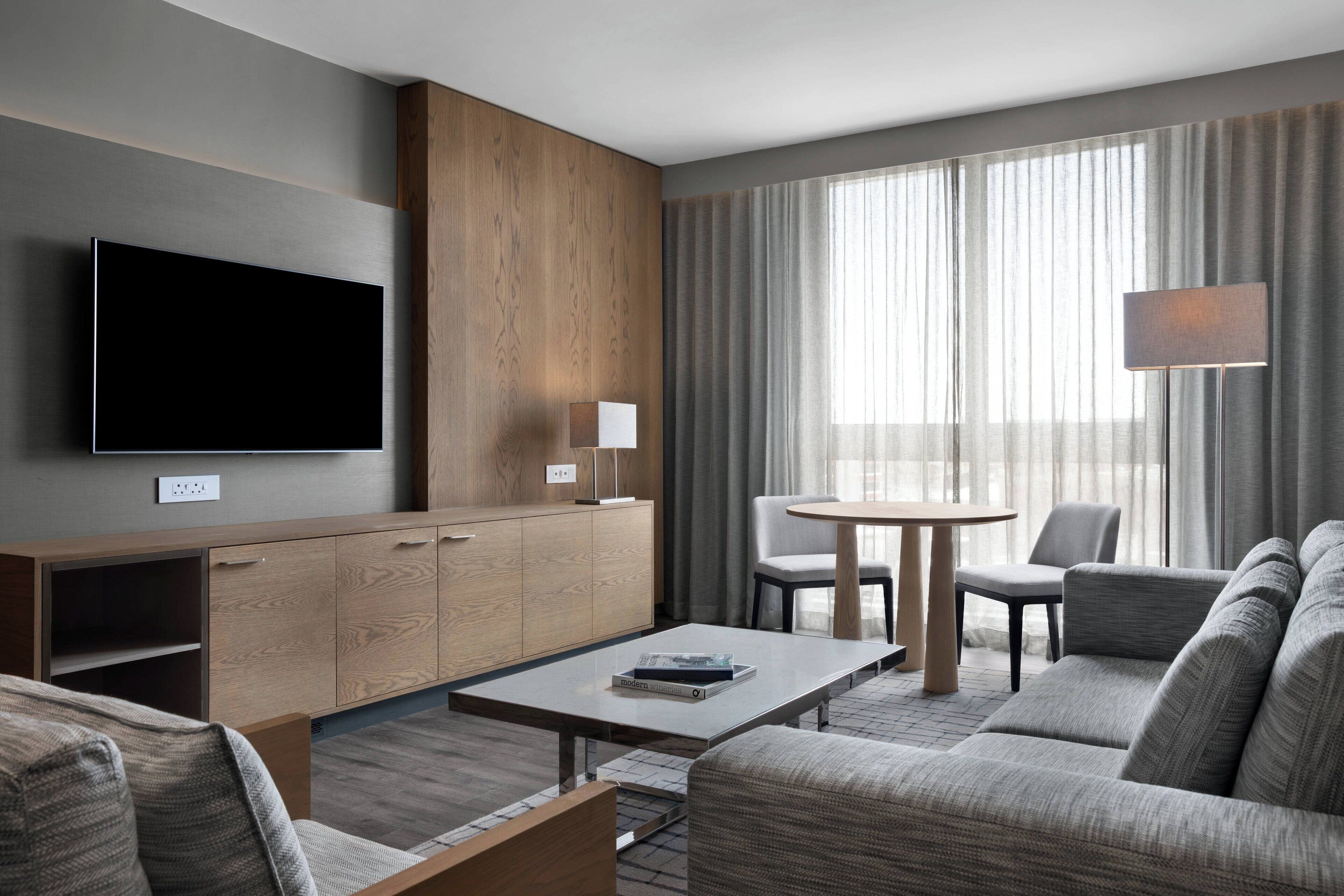 A modern hotel room with a flat-screen TV, wooden cabinet, grey sofa, round table, and large windows with curtains.