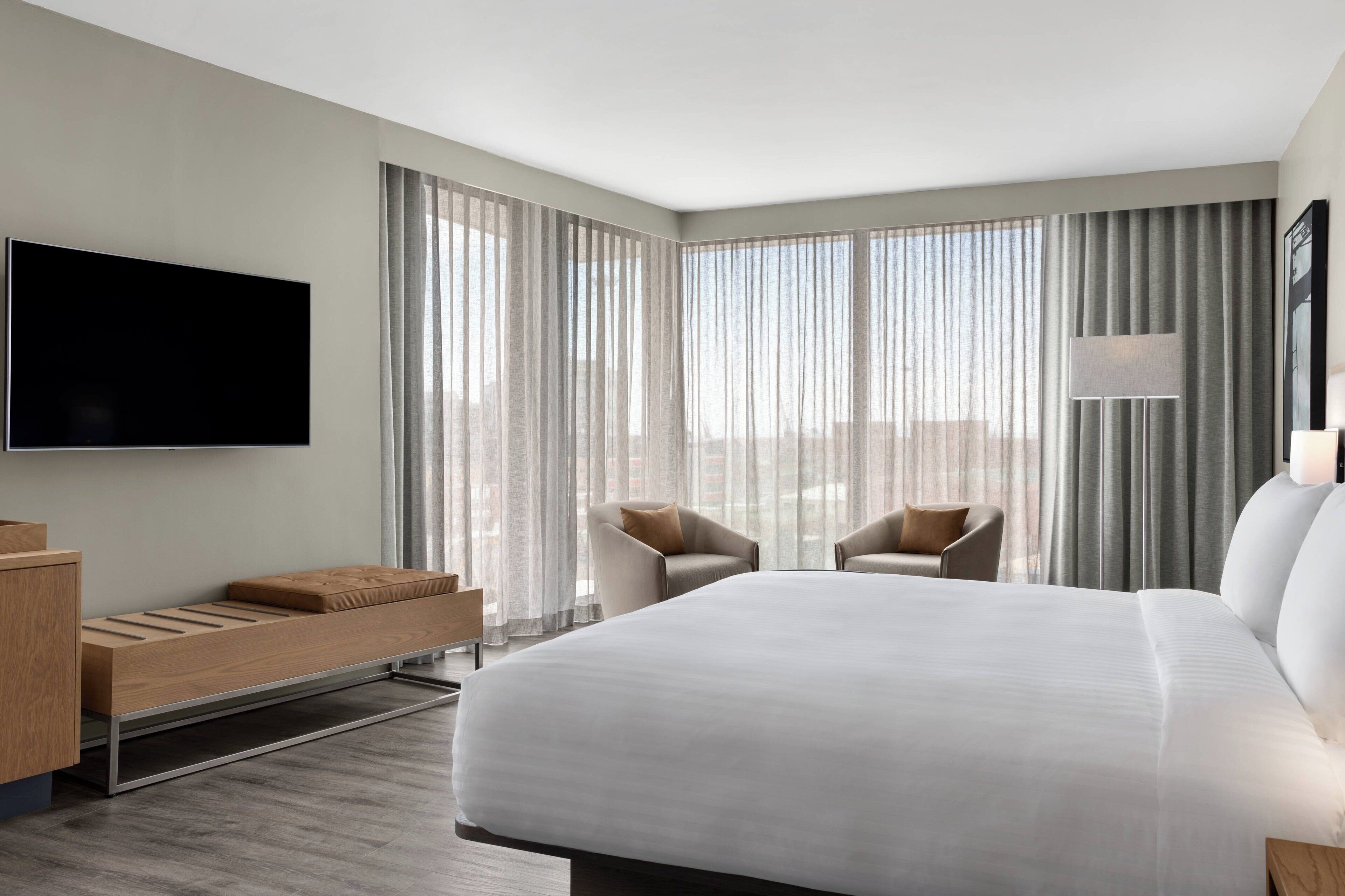 Quarto de hotel moderno com uma cama grande, duas poltronas, uma TV de tela plana e uma escrivaninha com cadeira.