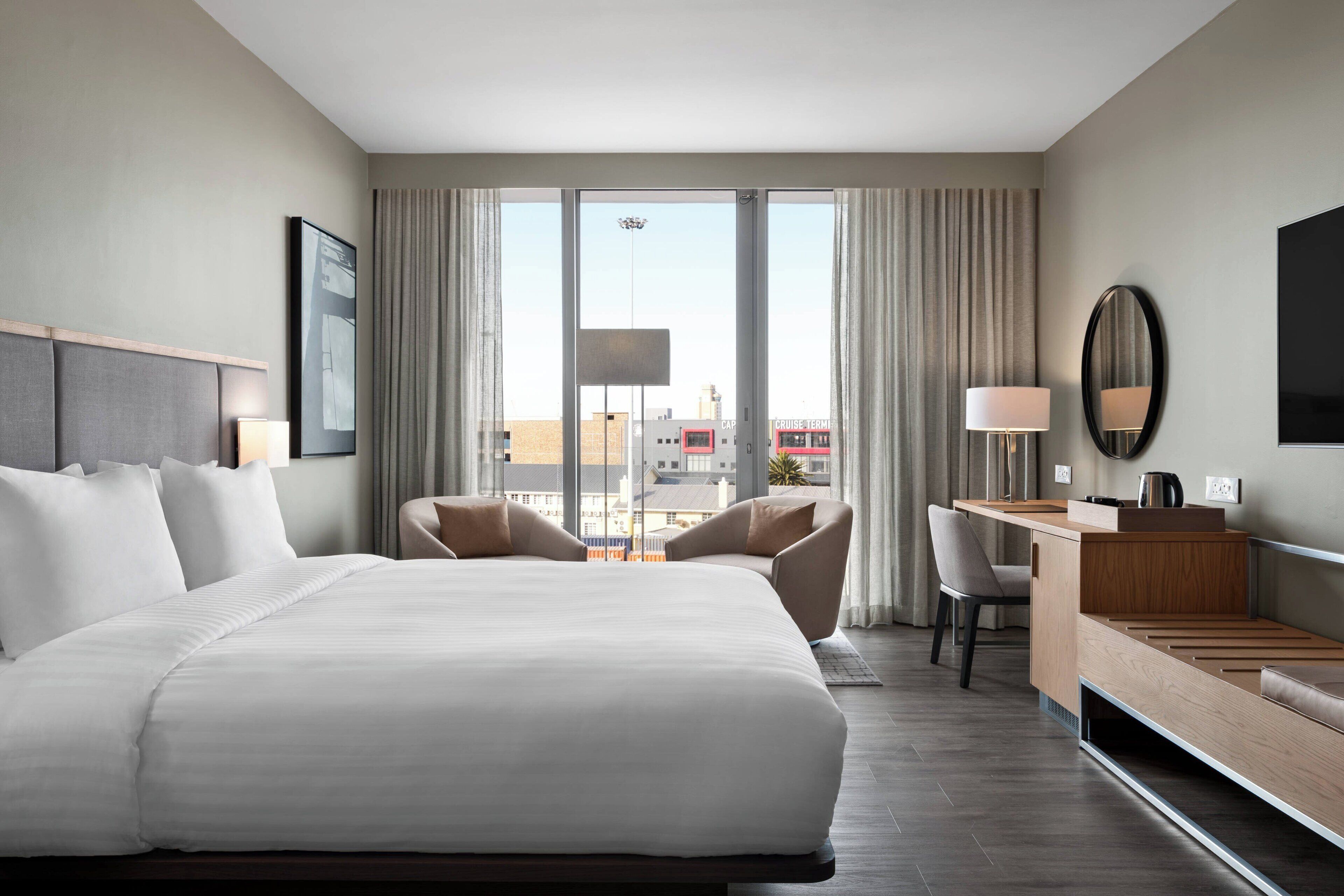 A hotel room with a large bed, a desk, a chair, and a view of the city through large windows.