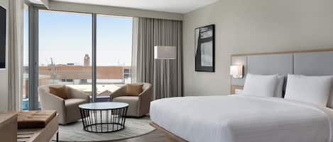 A modern hotel room with a large bed, a sofa, a small round table, and a view of the city through large windows.