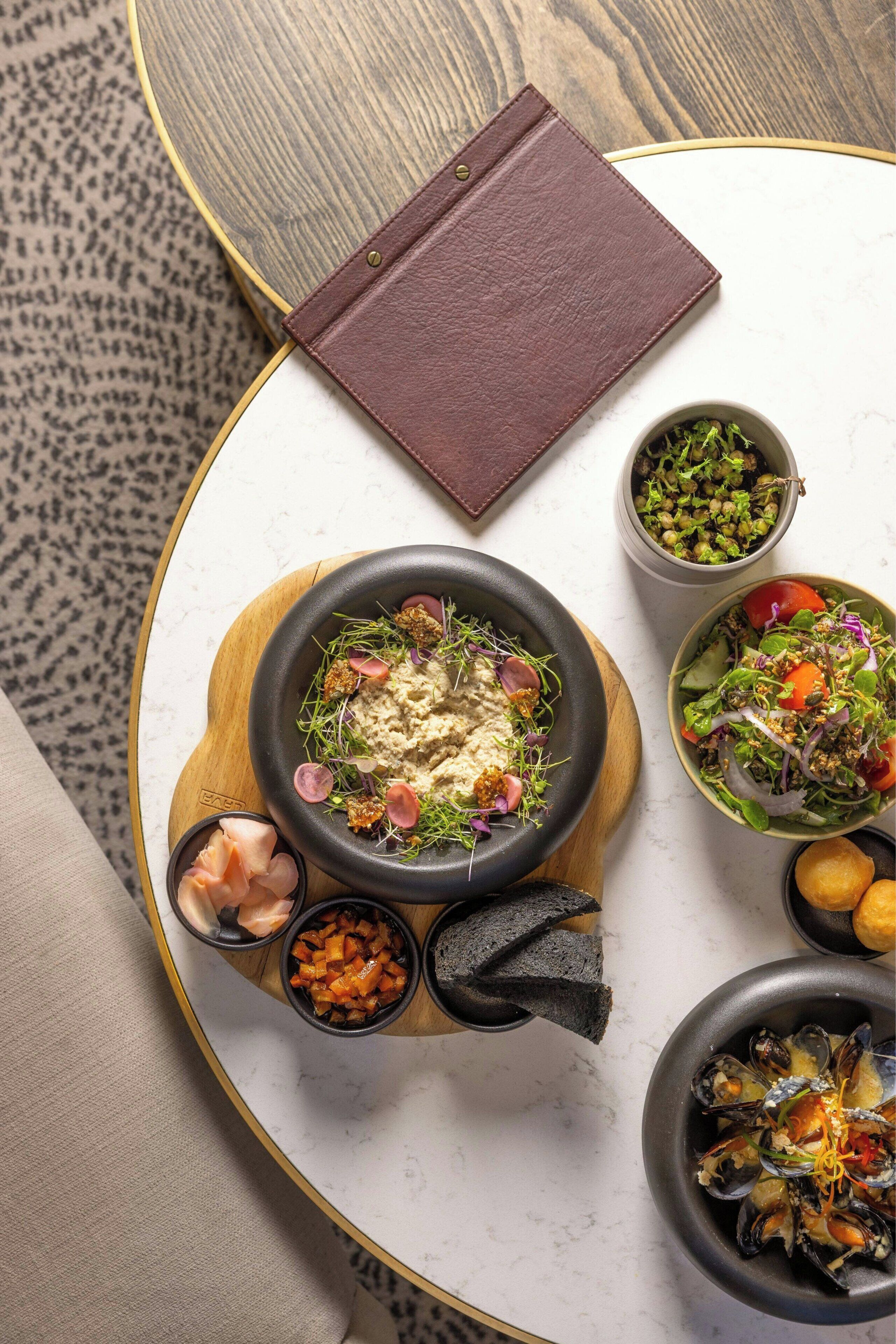 A dining table with a variety of dishes, including a bowl of hummus, a salad, and a seafood dish, along with a notebook and a cup of green vegetables.