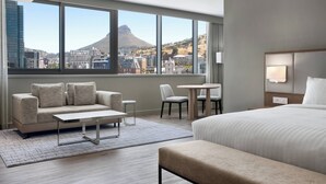 Quarto de hotel com uma janela grande, uma cama, um sofá, uma mesa de centro e uma pequena mesa de jantar.