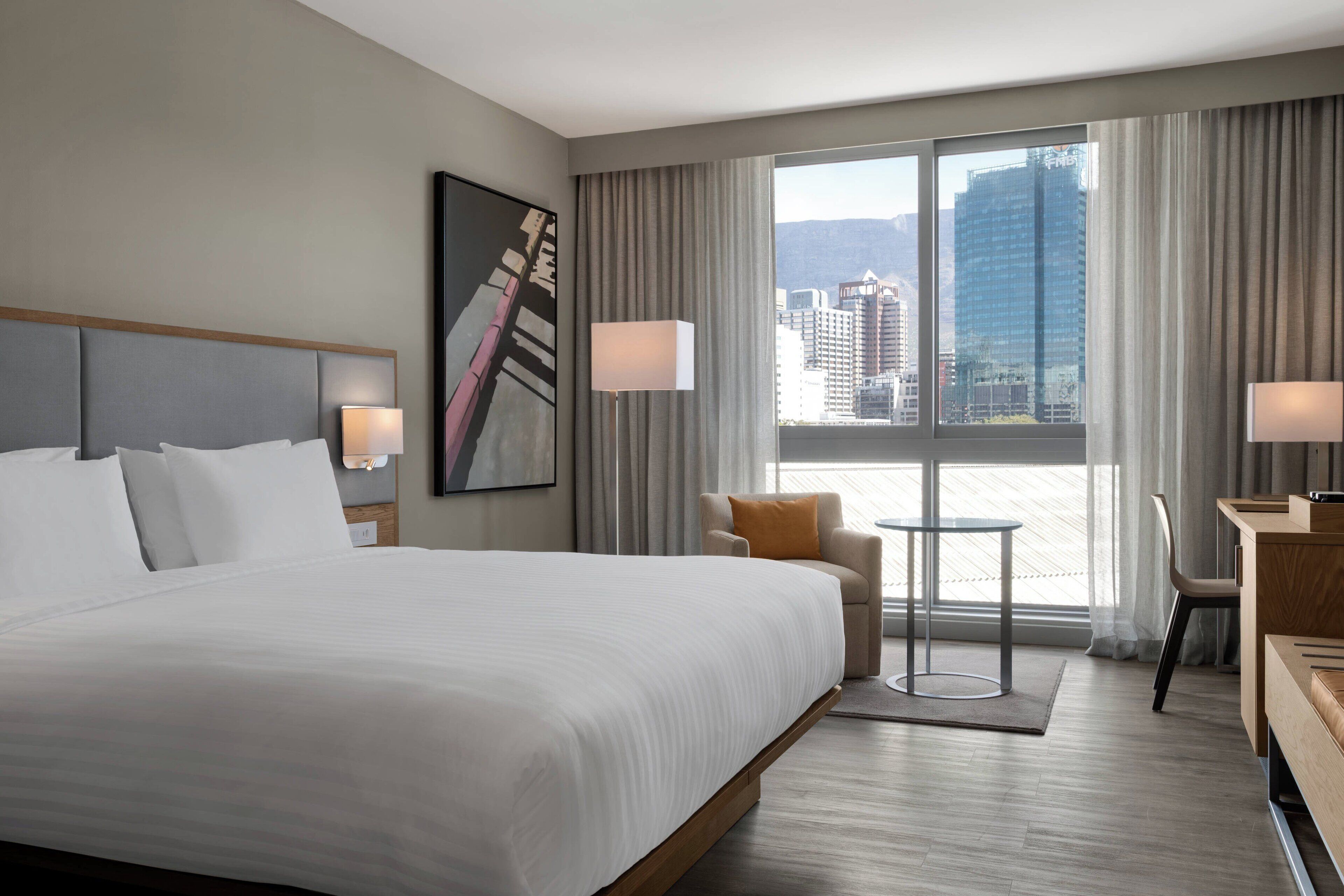 Quarto de hotel moderno com uma cama grande, uma escrivaninha, uma cadeira e vista para o horizonte urbano pelas janelas amplas.