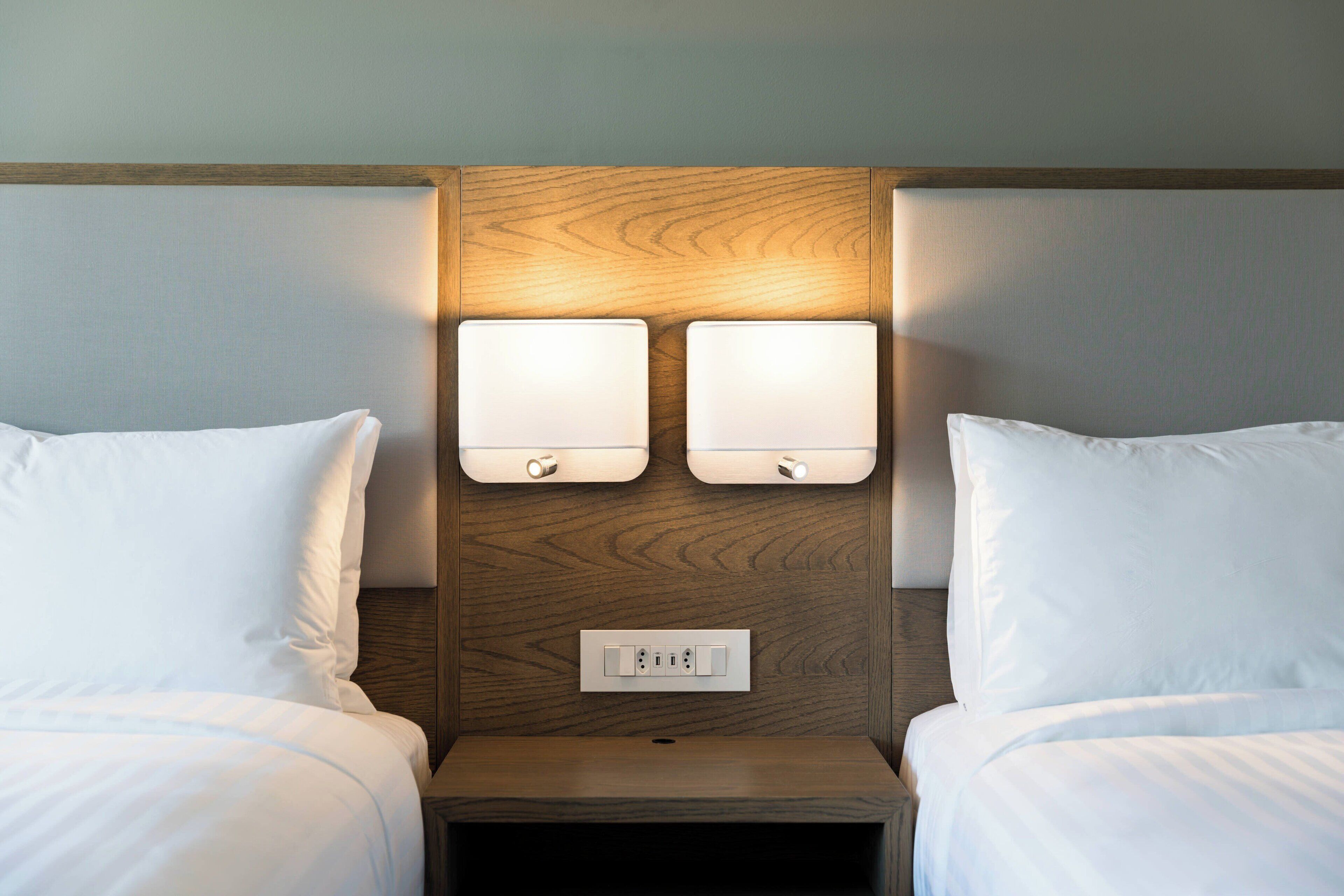 Une chambre d’hôtel avec deux lits, une tête de lit en bois et deux tables de chevet fixées au mur, chacune équipée d’une lampe.