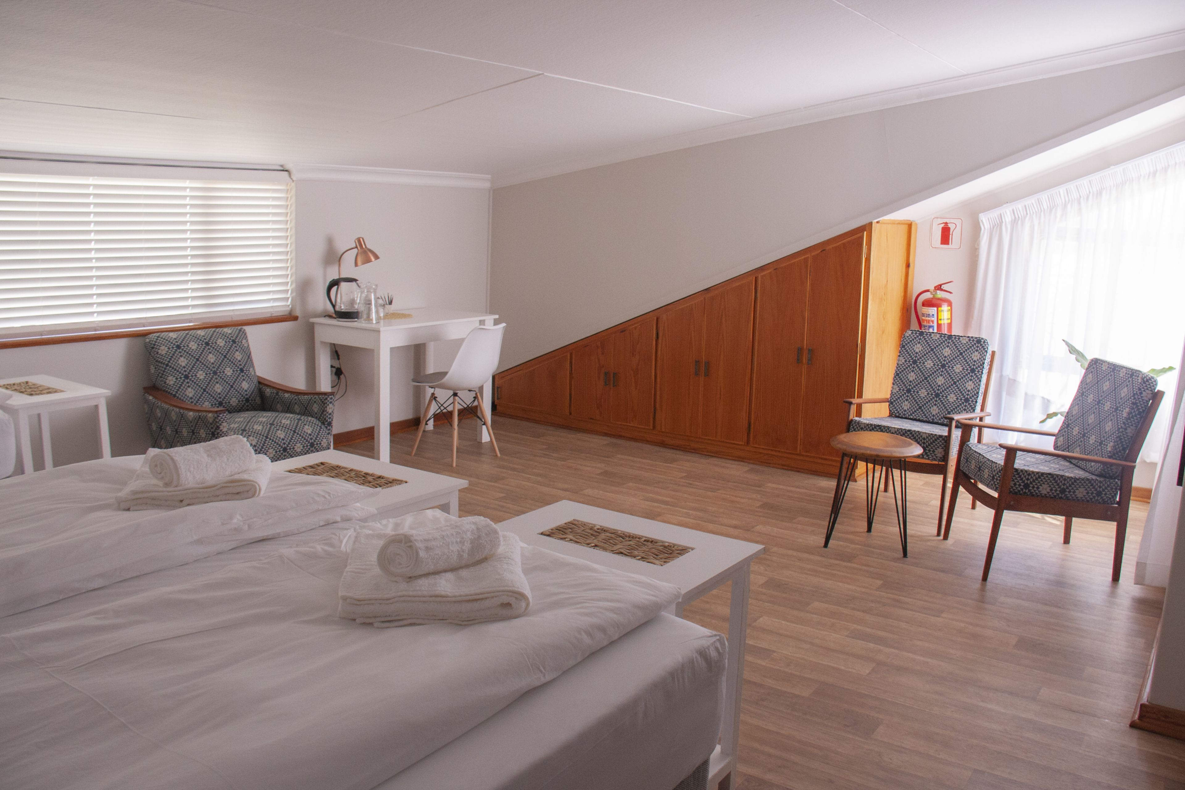 A hotel room with a large bed, two armchairs, a small table, and a desk. There is a window with blinds, a fire extinguisher on the wall, and a wooden wall panel.