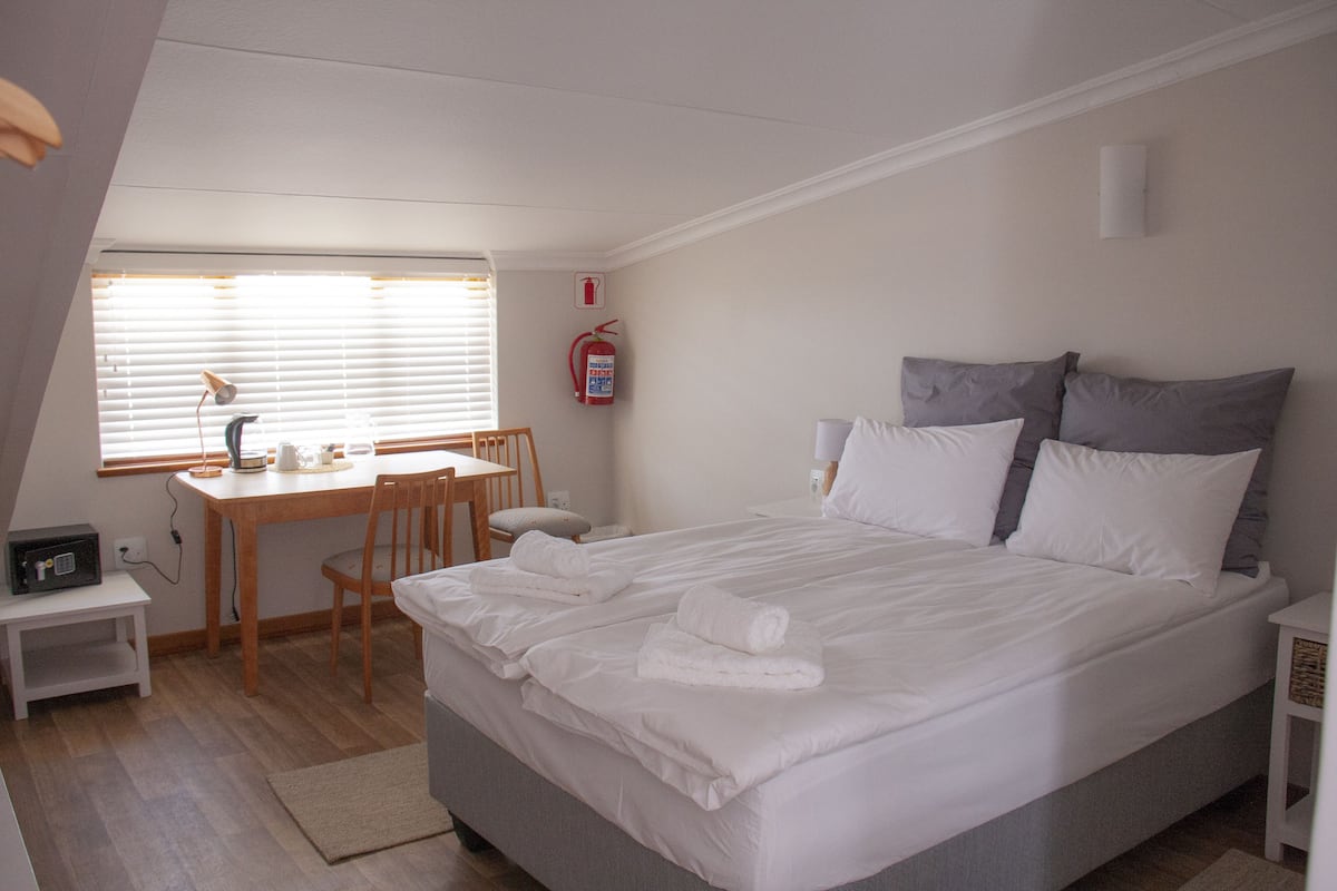 Een slaapkamer met een groot bed, een bureau en een stoel. Er hangt een brandblusser aan de muur en er is een raam met zonwering.