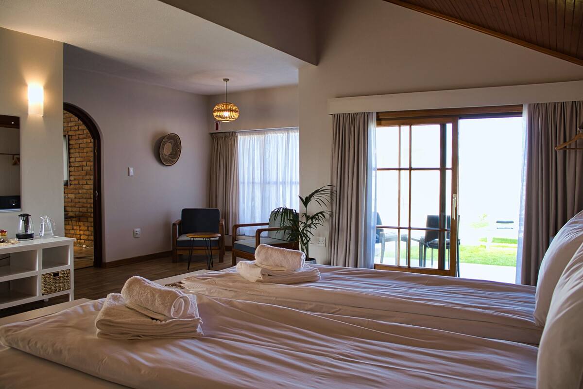 Een hotelkamer met een groot bed, een bureau met een lamp, een stoel, een plant en uitzicht op het landschap door een schuifdeur.