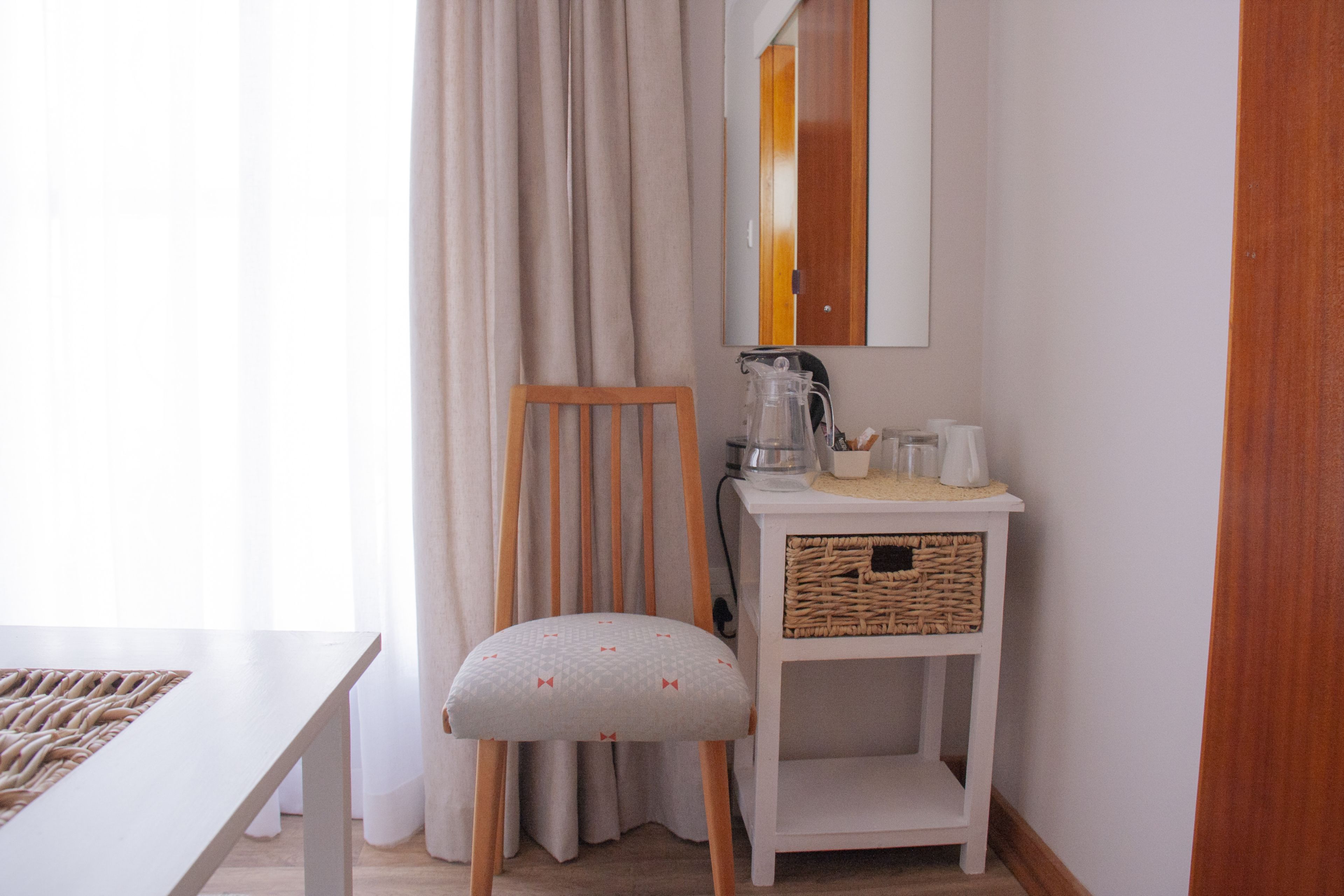 A small, neatly arranged room with a wooden chair, a white table, a white cabinet with wicker baskets, and a mirror on the wall.
