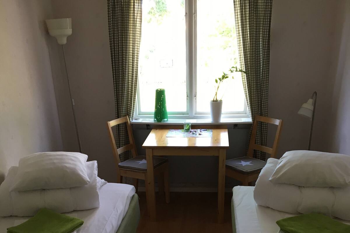 A room with two single beds, a small table with a chair, a window with curtains, and a lamp.