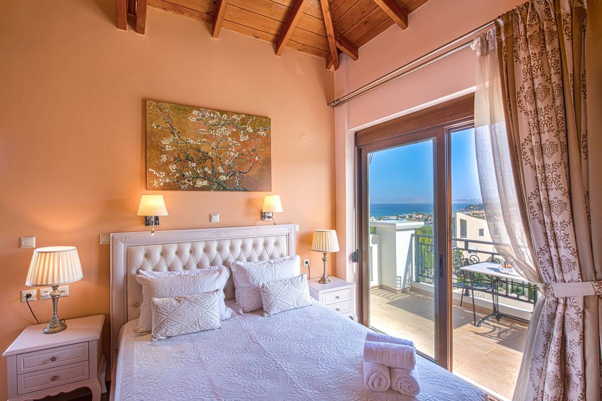A bedroom with a large bed, two bedside lamps, a painting on the wall, and a balcony with a view.