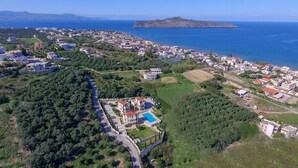 Vista aérea de un pueblo costero con un complejo de villas destacado, que incluye una piscina, rodeado de abundante vegetación y edificios residenciales.