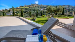 Un espacio al aire libre iluminado por el sol con sillones de descanso, una mesa con un libro, vasos y una bebida, con vistas a una ladera pintoresca con edificios.