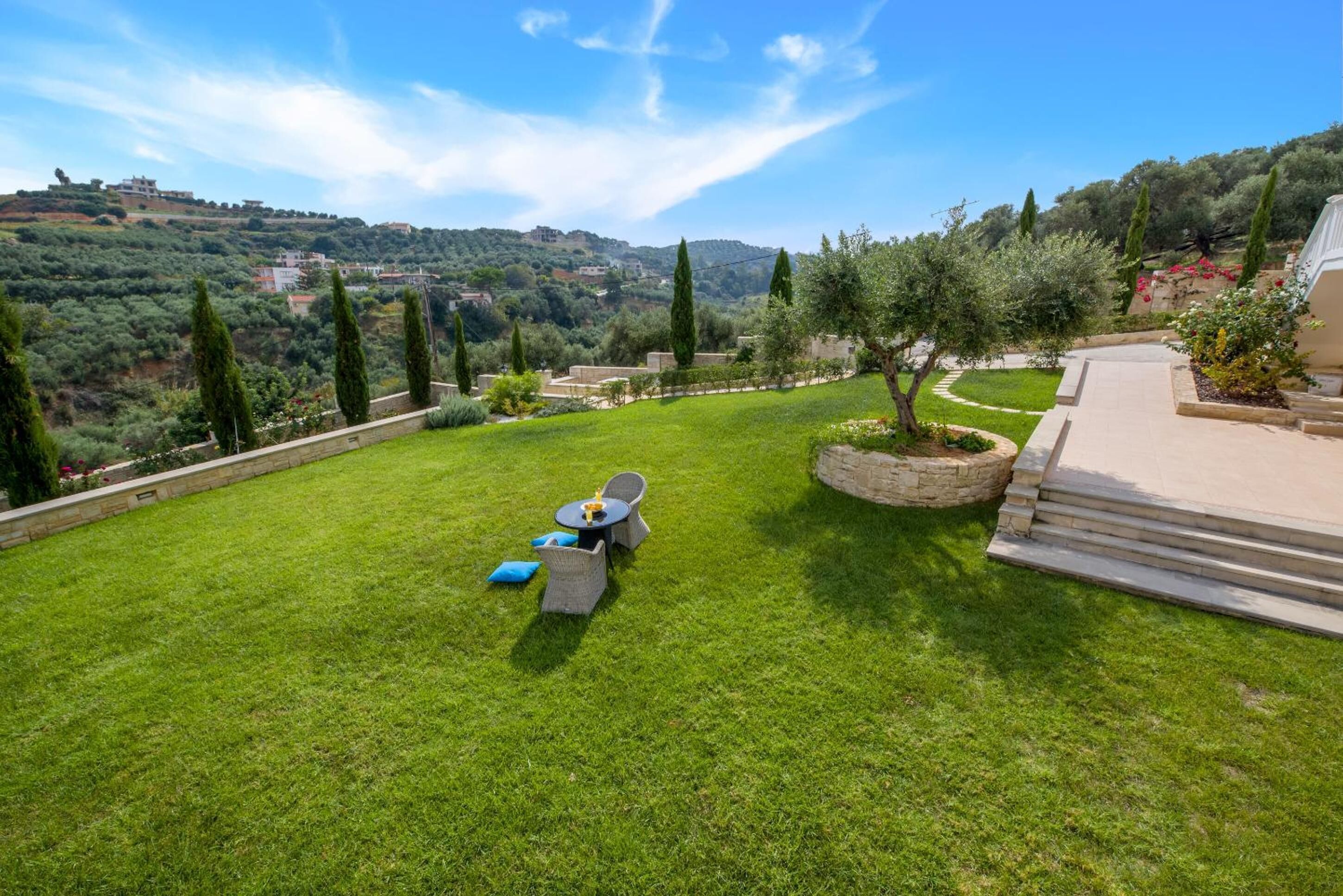 A well-maintained lawn with a seating area, a stone pathway, and a view of a hilly landscape with buildings.
