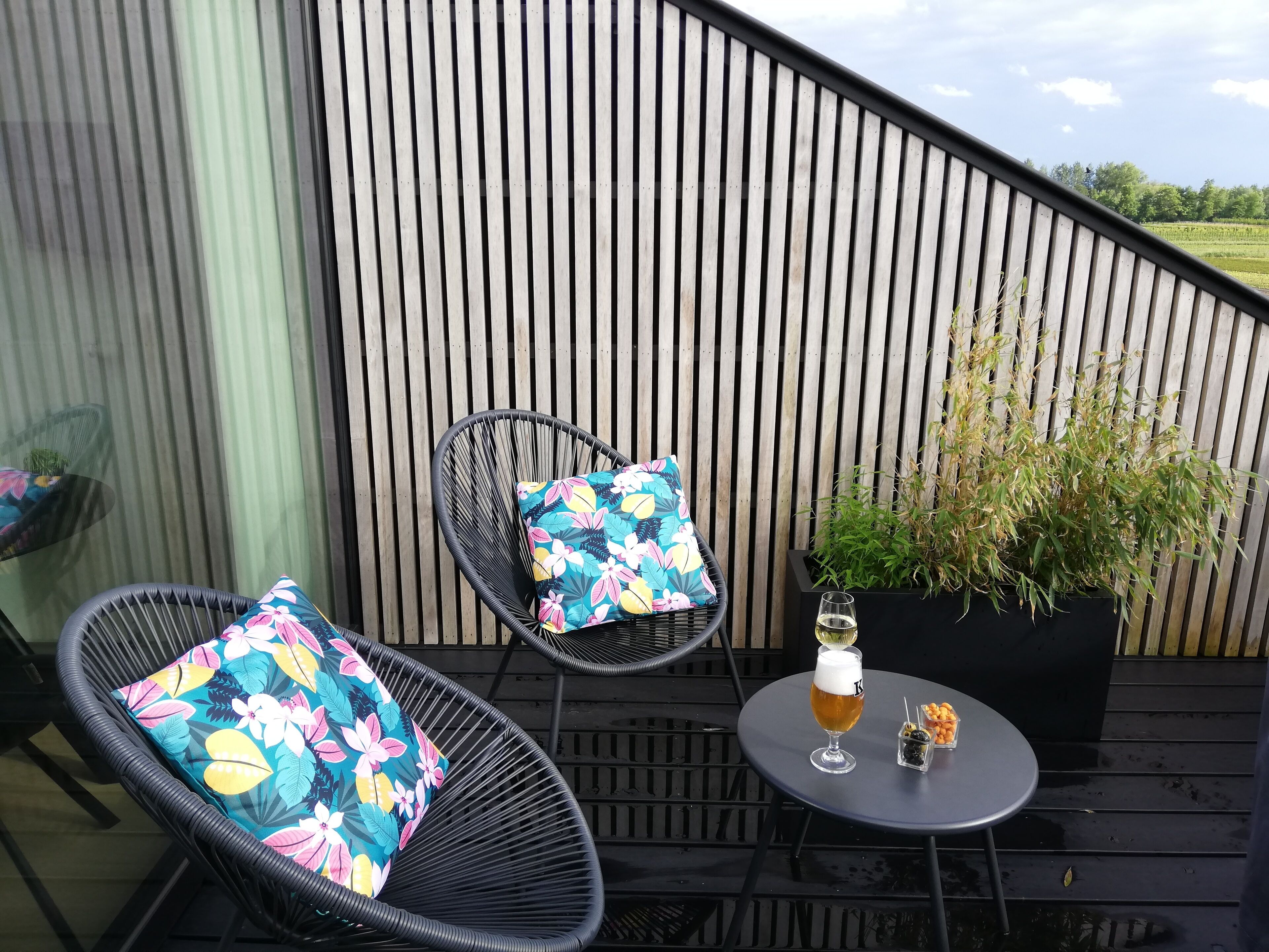 A patio area with two wicker chairs, one with a floral cushion, and a small round table with a glass of beer and snacks.