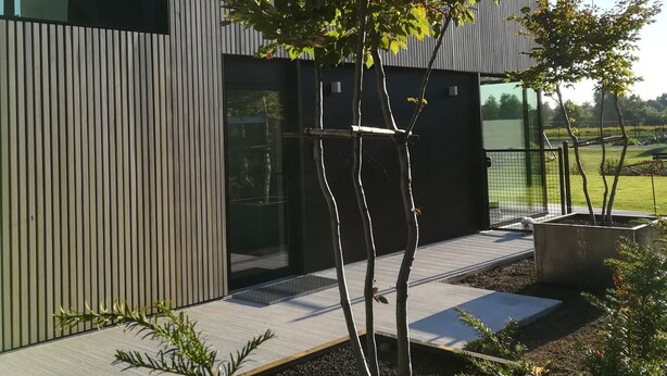Een modern gebouw met verticale beplating, een groot raam en een aangelegde tuin met jonge bomen en struiken.