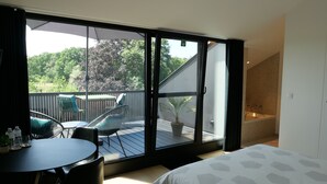 Een slaapkamer met een grote schuifdeur die leidt naar een balkon met buitenmeubilair en uitzicht op bomen.