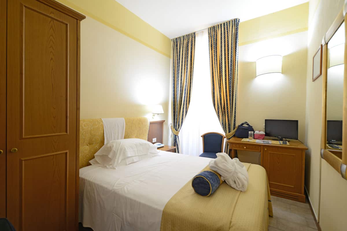 Ein Hotelzimmer mit einem Bett, einem Schreibtisch mit Computer, einem Stuhl, einer Lampe und Vorhängen.