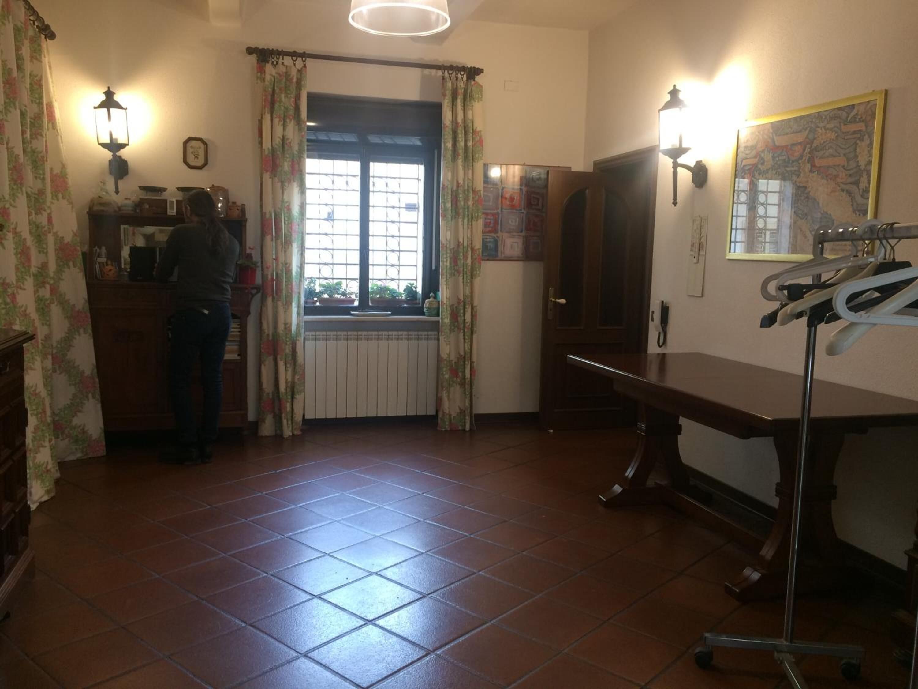 Une pièce avec un sol carrelé, un bureau avec une chaise, un radiateur, une fenêtre avec des rideaux et des lampes fixées au mur.