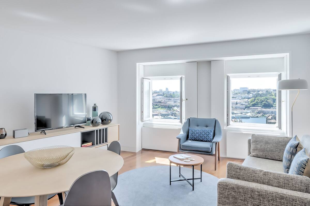 A modern living room with a flat-screen TV, a dining table, and a sofa. There are two windows with a view of a cityscape.