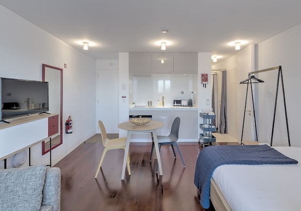 A modern hotel room with a bed, a TV, a dining table with chairs, a kitchen area, and a wardrobe.
