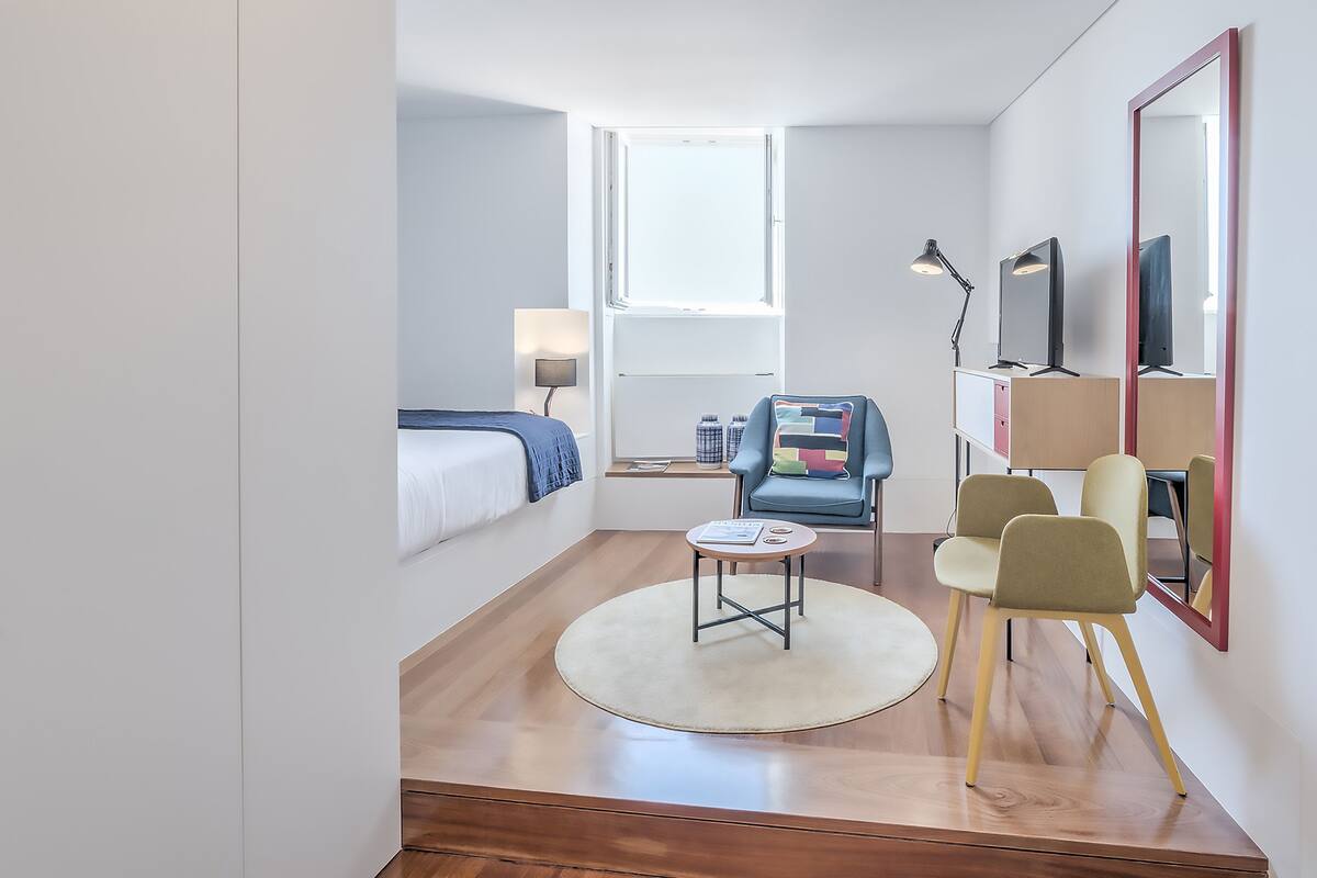 A compact hotel room with a bed, a small round table, a chair, a desk with a computer, and a large mirror.