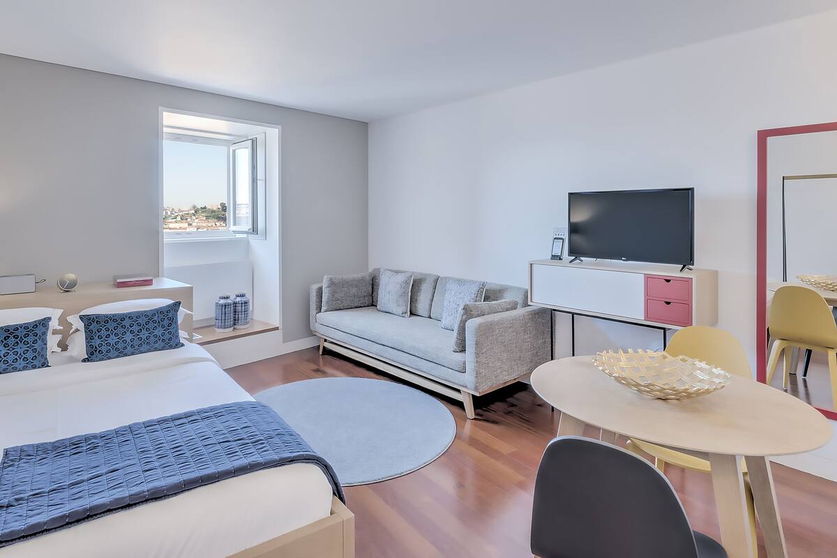 A modern hotel room with a bed, sofa, TV, and dining area.