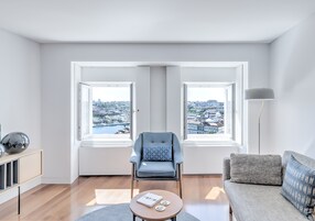 Una sala moderna con vistas a un paisaje urbano a través de amplios ventanales, un sillón gris, un sofá gris y una pequeña mesa redonda con un libro y dos objetos decorativos.
