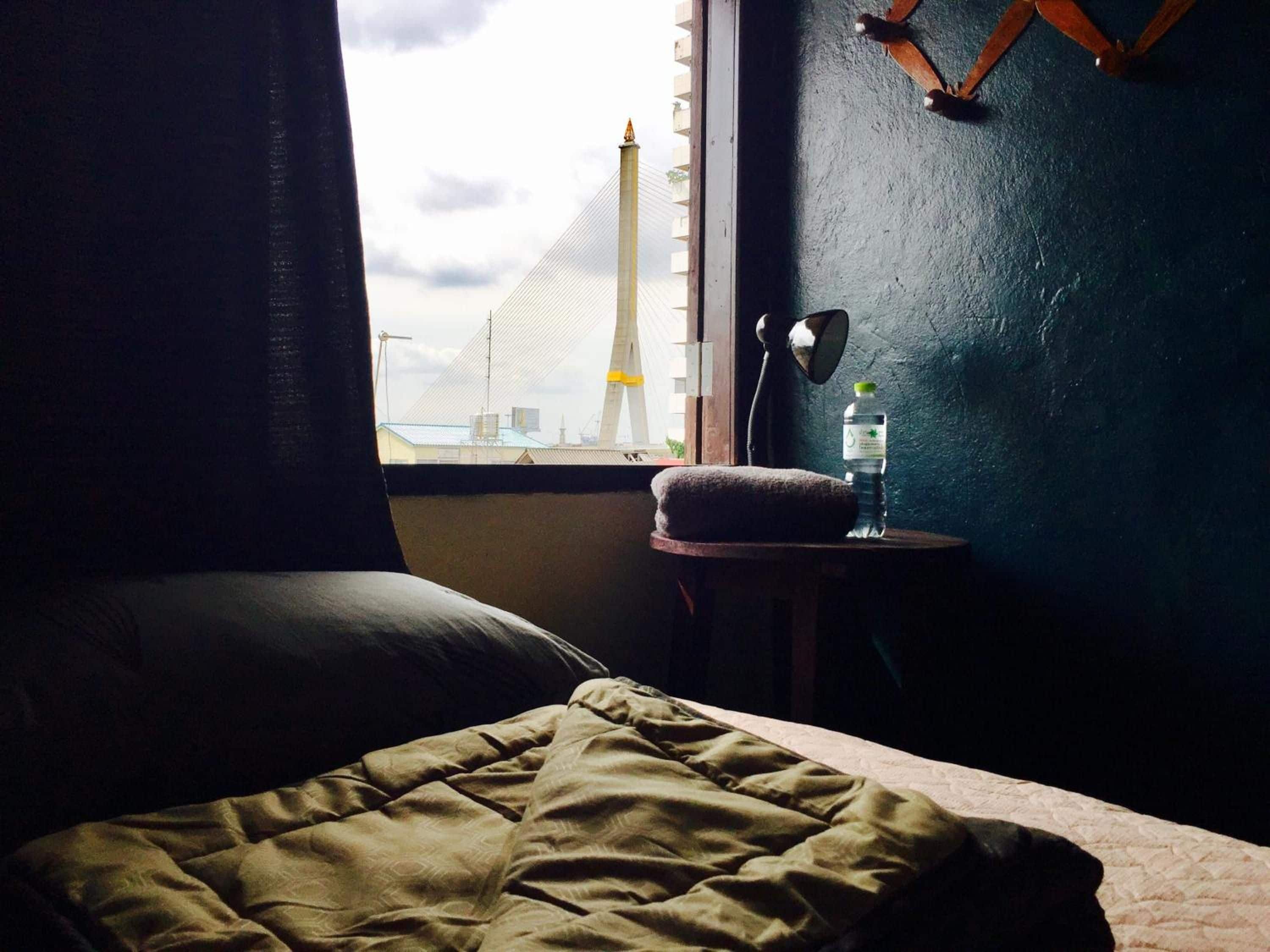 A bed with a green blanket, a small bedside table with a water bottle and a lamp, and a view of a bridge and tower through a window.