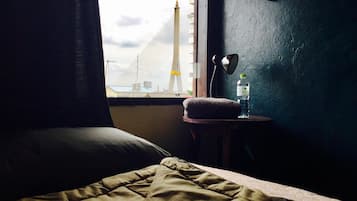A bed with a green blanket, a small bedside table with a water bottle and a lamp, and a view of a bridge and tower through a window.