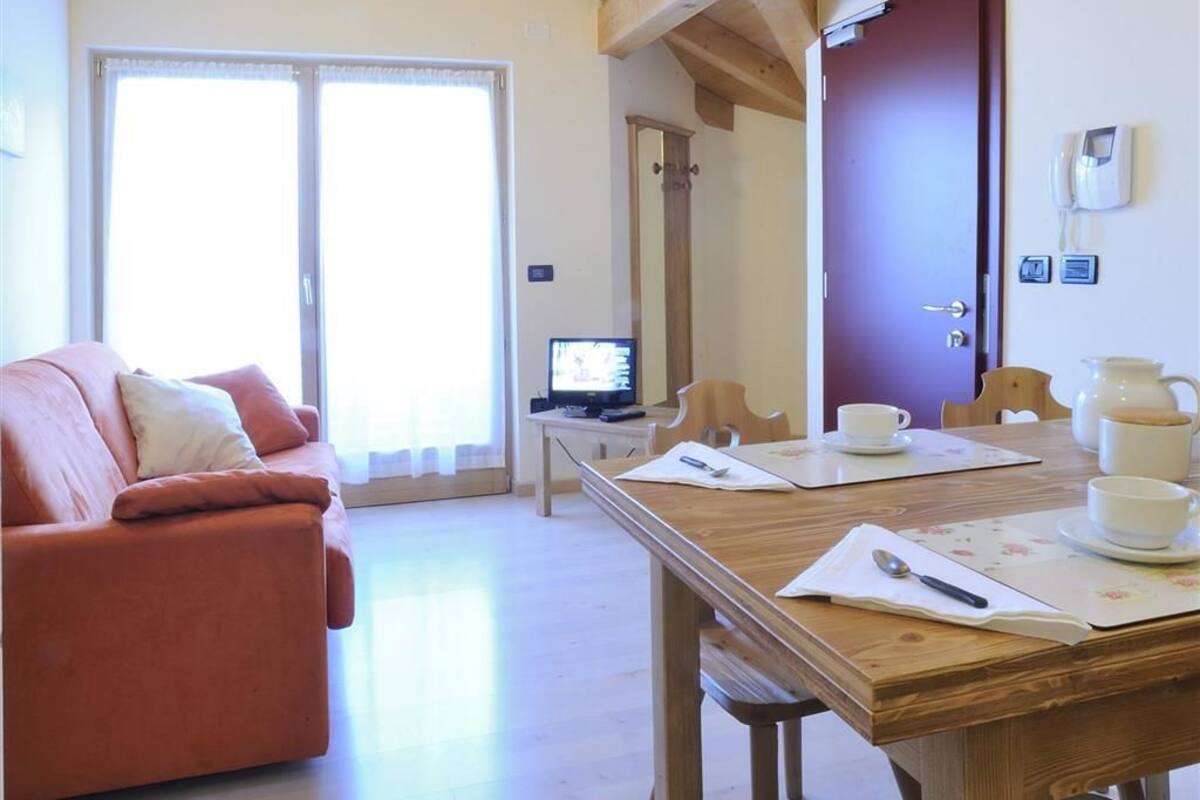 A room with a wooden ceiling, a sofa, a dining table with chairs, a television, and a door.