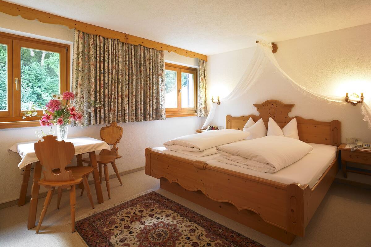 A bedroom with a wooden bed, a nightstand, a bedside table, a chair, a desk with a chair, a window with curtains, and a vase with flowers.