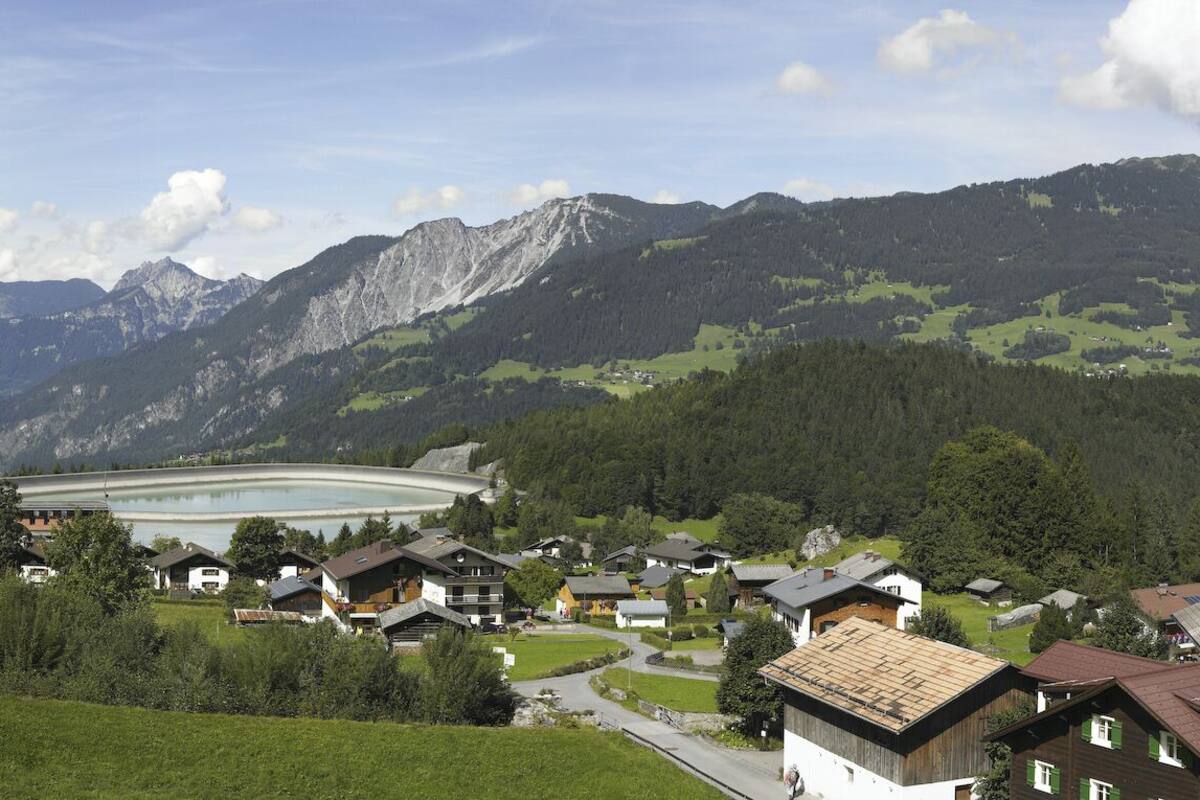 A mountain village with traditional houses, a large building possibly a hotel, and a valley with dense forests and mountains in the background.