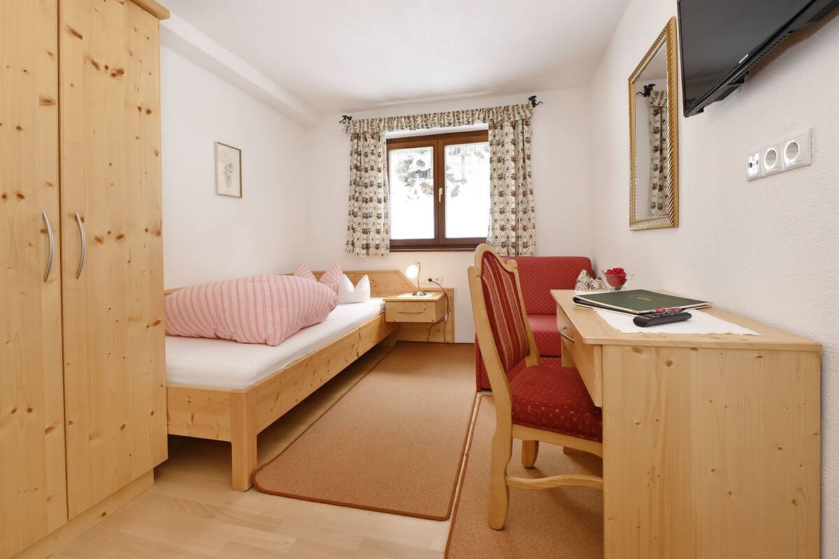 A small, cozy room with a single bed, a desk with a chair, a TV, and a window with curtains.