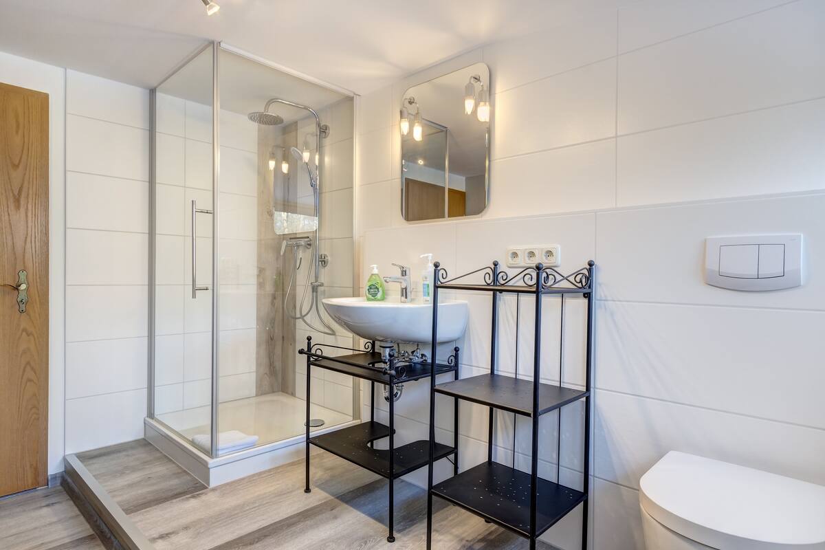 Un baño moderno con ducha de vidrio, un lavabo blanco y una repisa metálica negra, y un inodoro blanco.