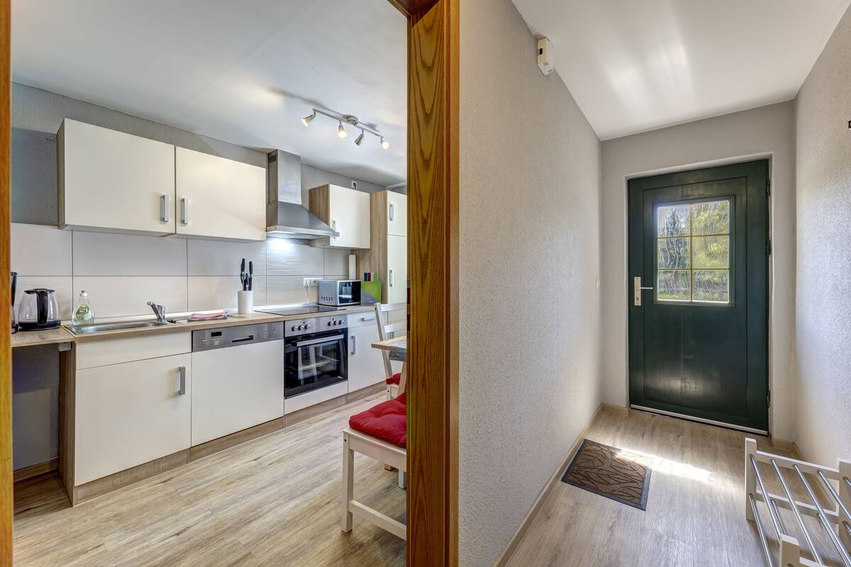 Una cocina moderna con armarios blancos, lavavajillas y estufa. Hay una silla roja y una puerta con ventana.