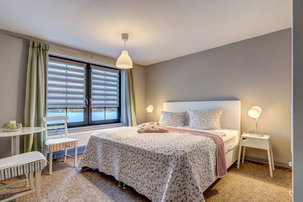Un dormitorio con cama, mesitas de noche, una silla, ventana con persianas y lámpara colgante.