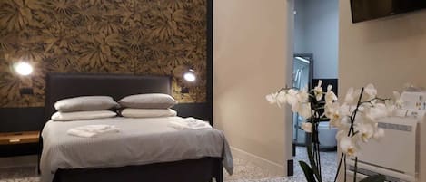 A modern bedroom with a large bed, a dark headboard, and a wall with a leafy pattern. There is a white orchid plant and a TV mounted on the wall.