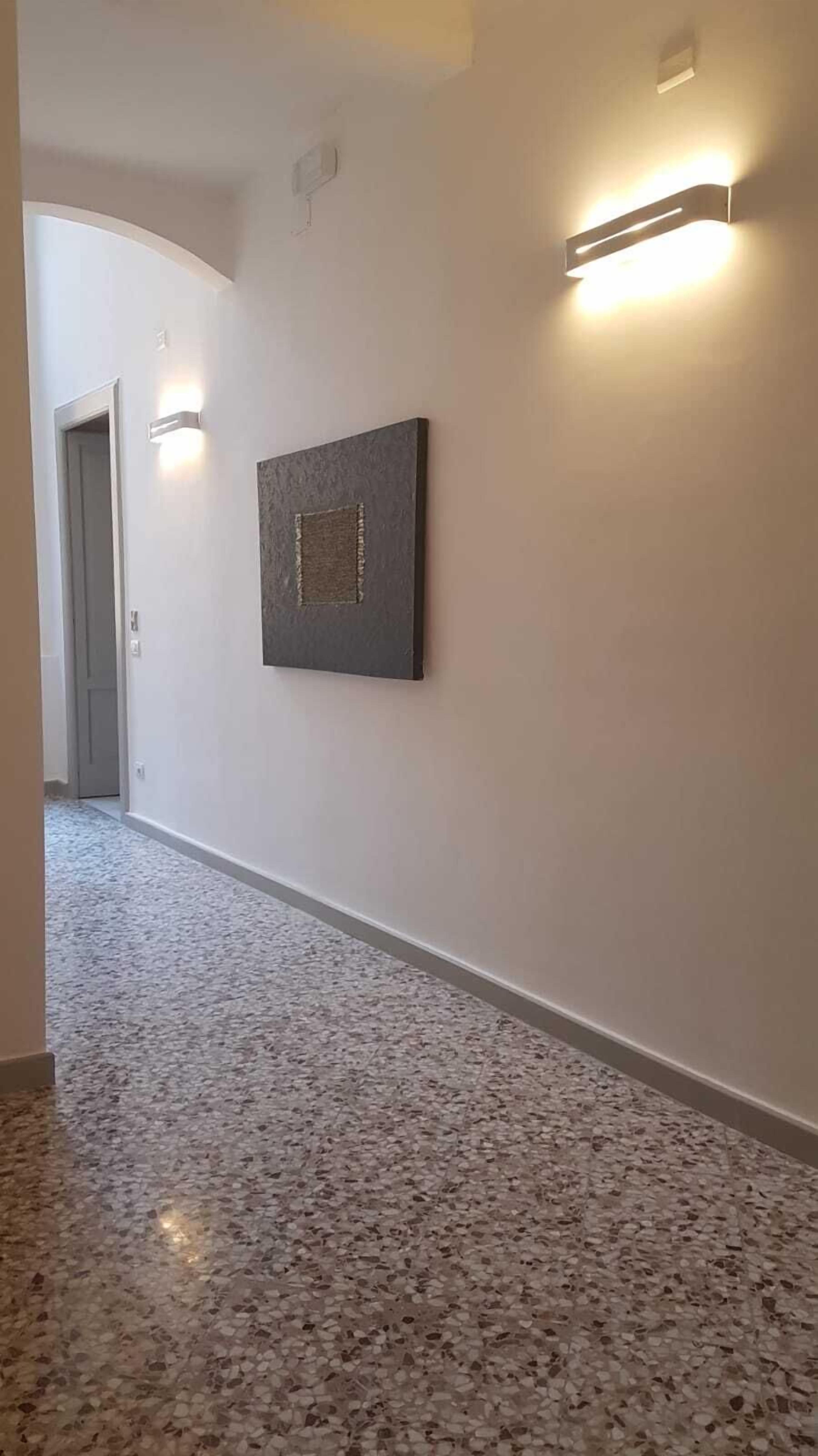 A hallway with a terrazzo floor, a framed artwork on the wall, and two wall-mounted light fixtures.