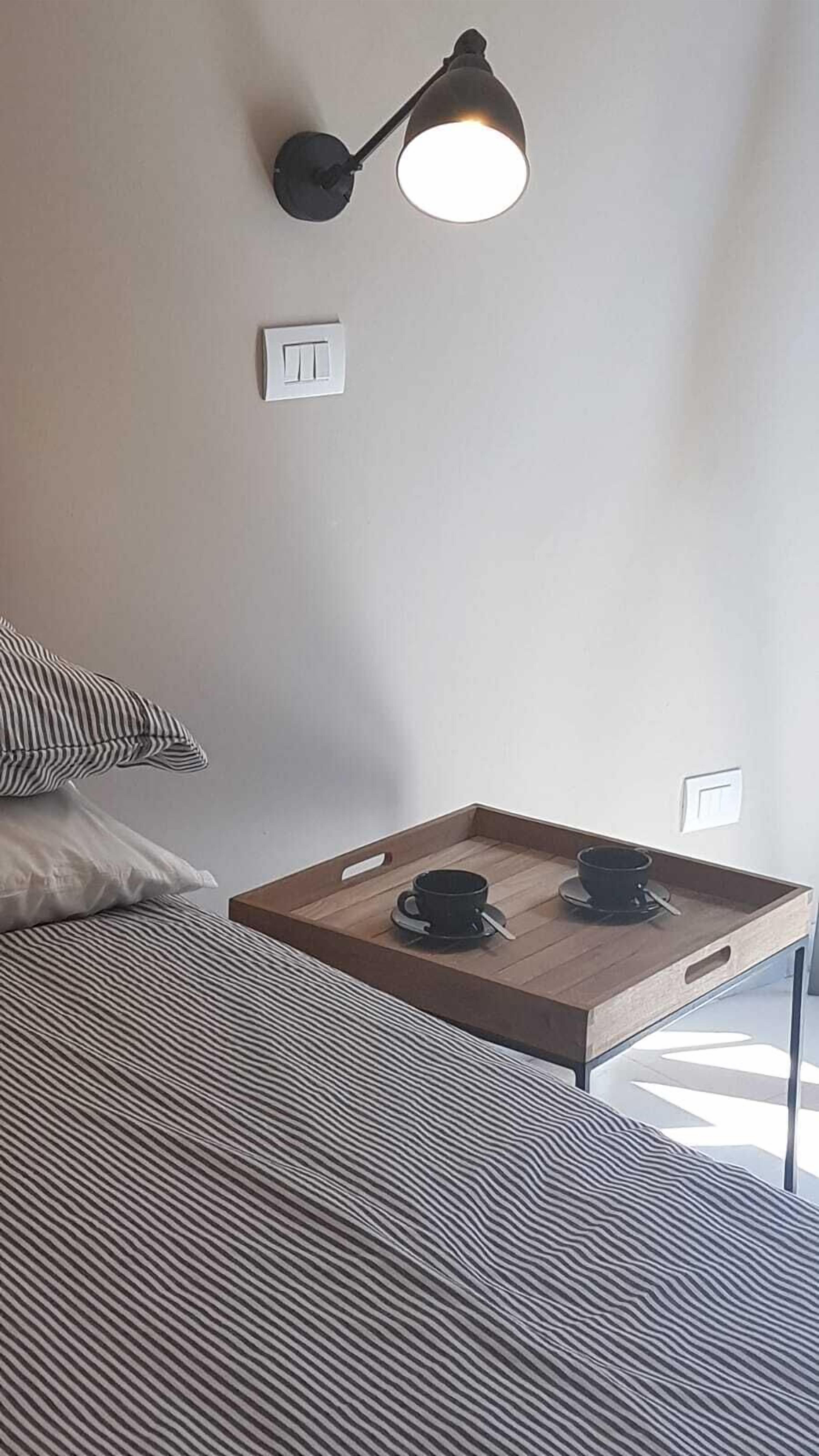 A neatly arranged bedroom with a bed, a bedside table with a cup and saucer, a lamp, and a power outlet on the wall.