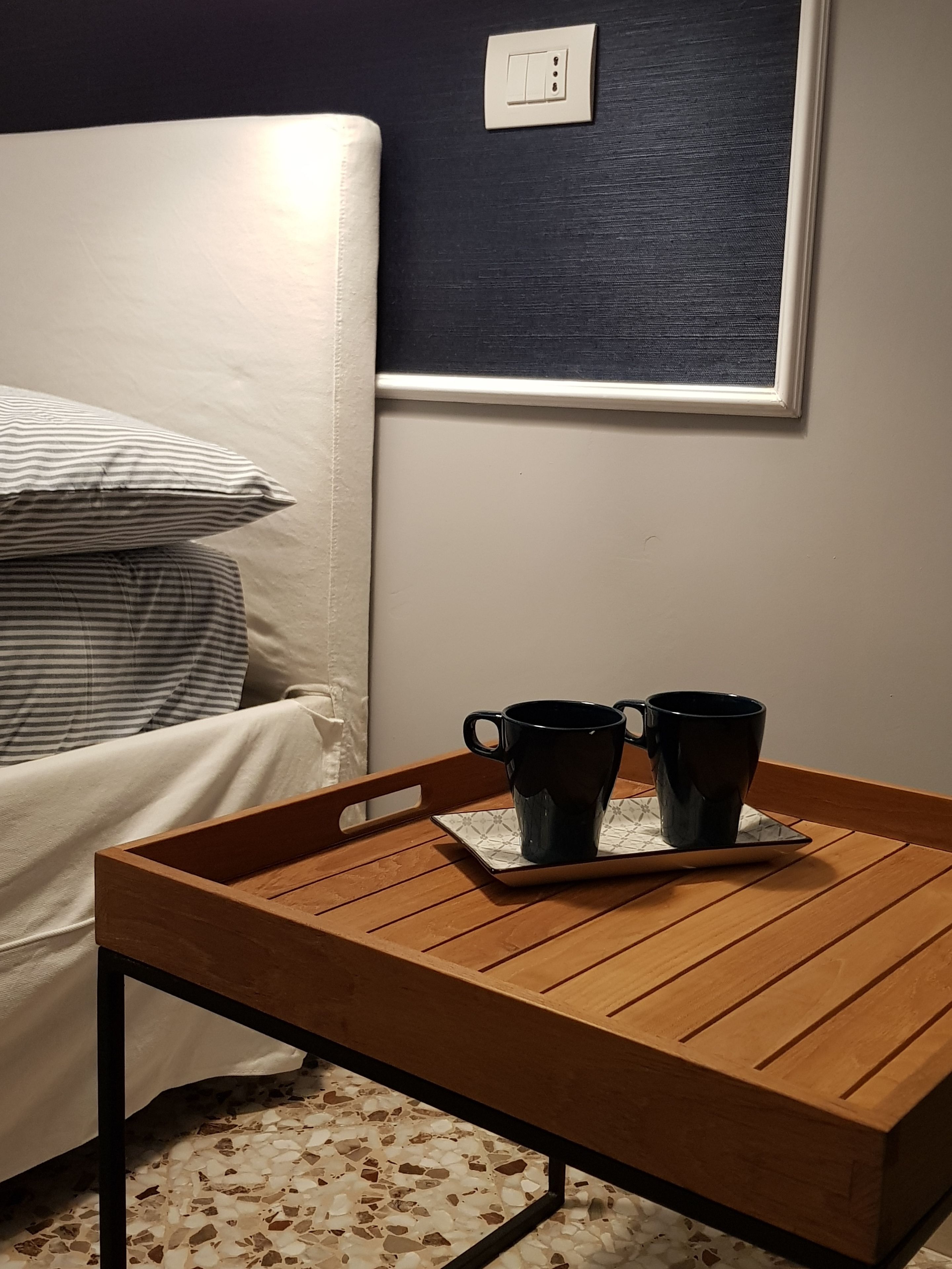 Una mesita de madera con dos tazas negras sobre una bandeja, una cama con almohadones a rayas y un enchufe de pared.